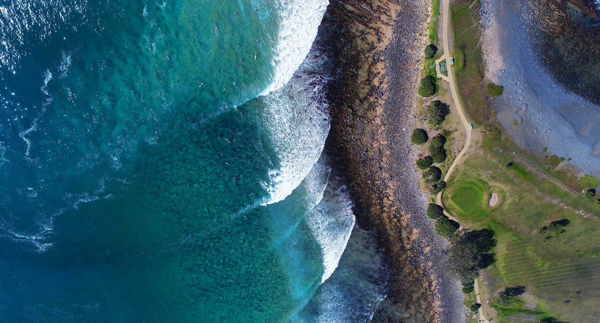 Aerial view of ocean waves crashing on a rocky shoreline with adjacent green park area and walking paths.