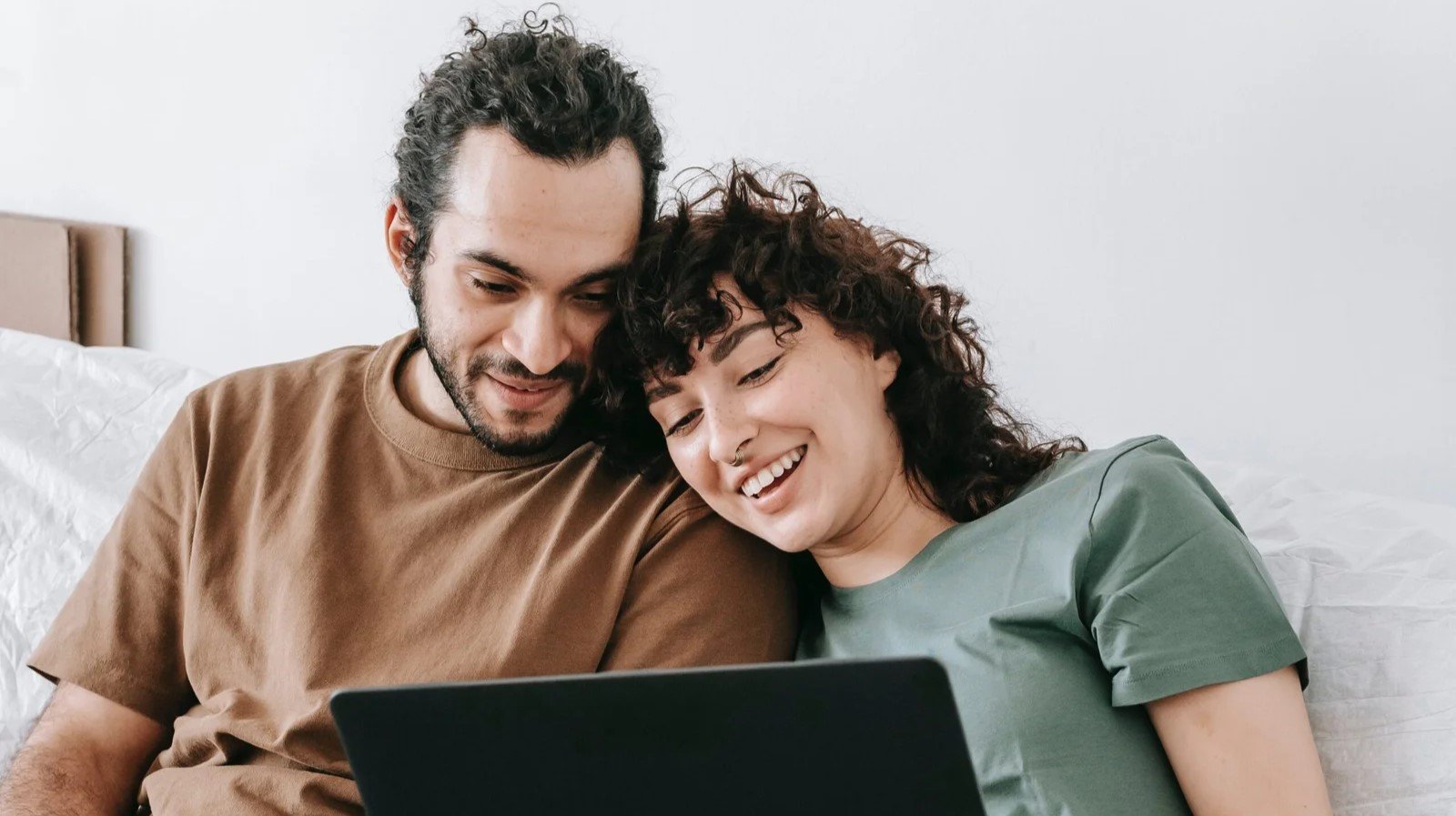 A couple sits on a couch, smiling and looking at a laptop together.