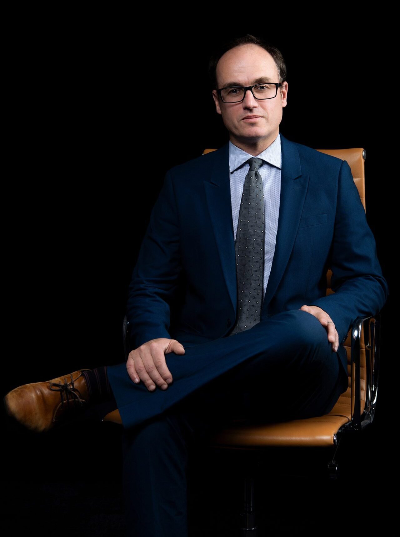 Iain Mark sits in an office chair, wearing a suit and glasses.