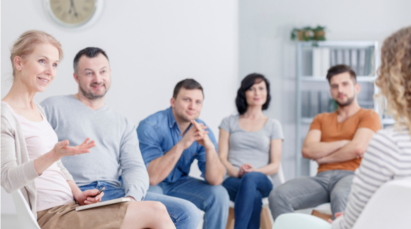 A group of six people in a discussion setting, potentially a family council, seated in a circle with a clock in the background.