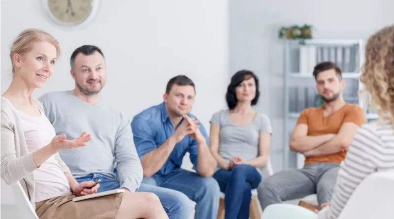 A group of six people in a discussion setting, potentially a family council, seated in a circle with a clock in the background.