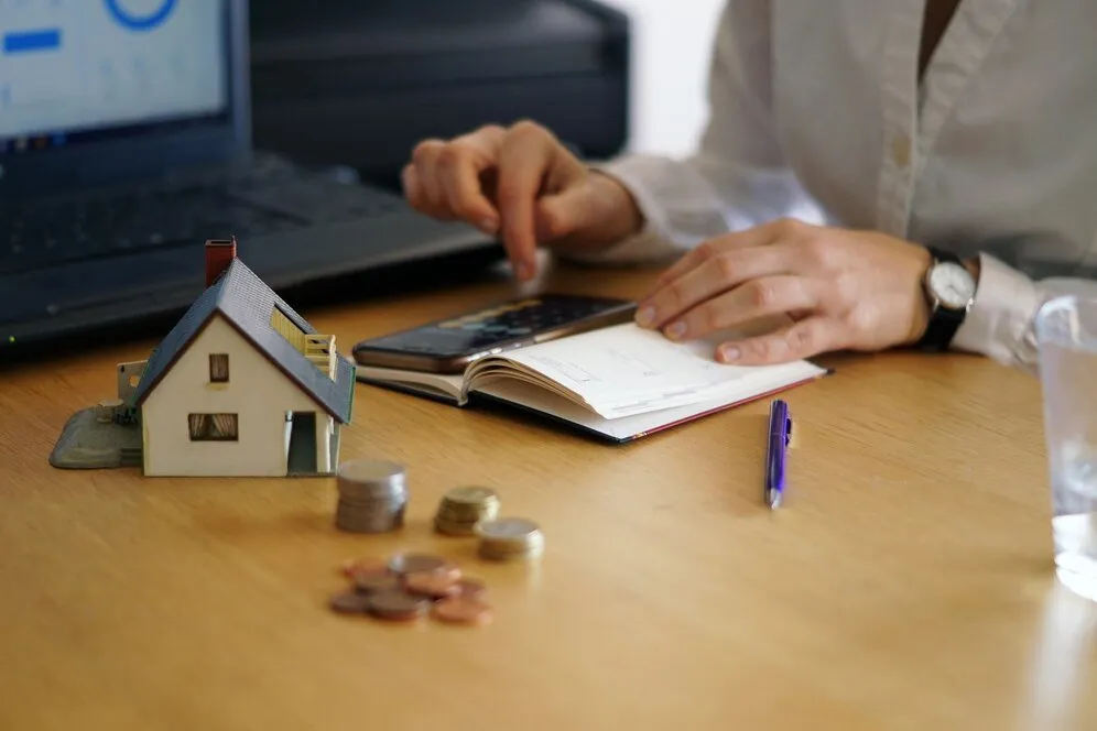Woman reviewing finances, house model & coins on desk. High net-worth family financial planning.