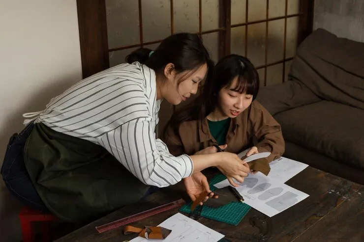 Two women collaborate on a leather crafting project, studying patterns and materials.