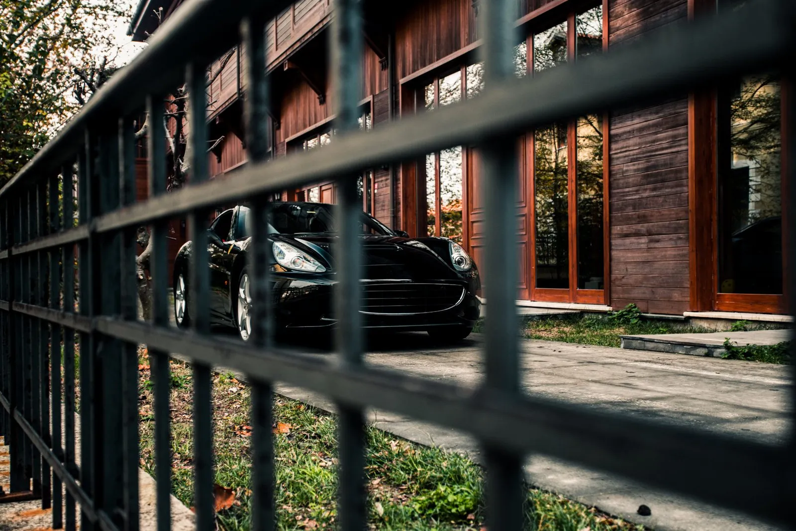 Black Ferrari parked in front of a brown wooden house, viewed through a metal fence.