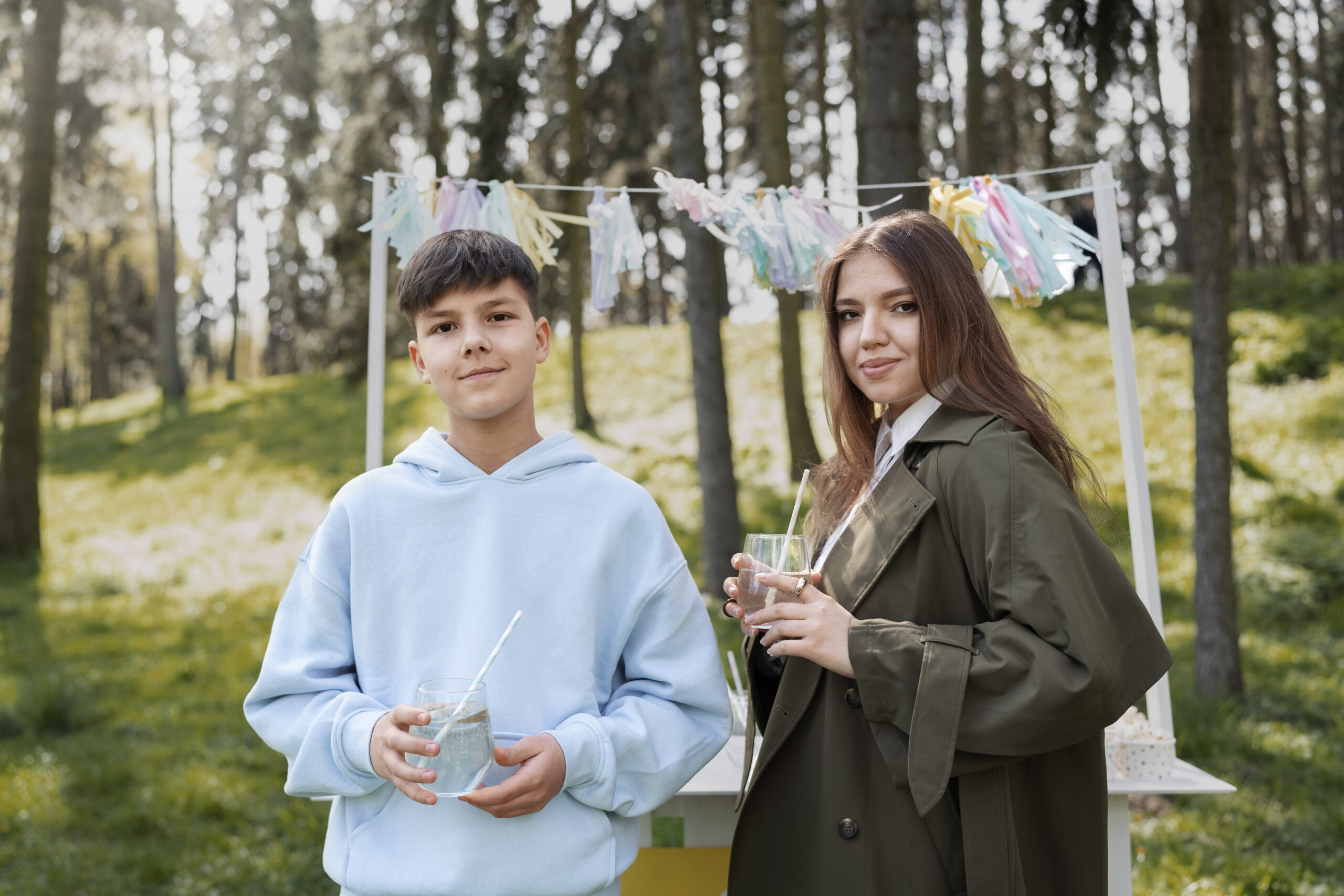 Two young people, a boy and a girl, stand at a lemonade stand in a park.