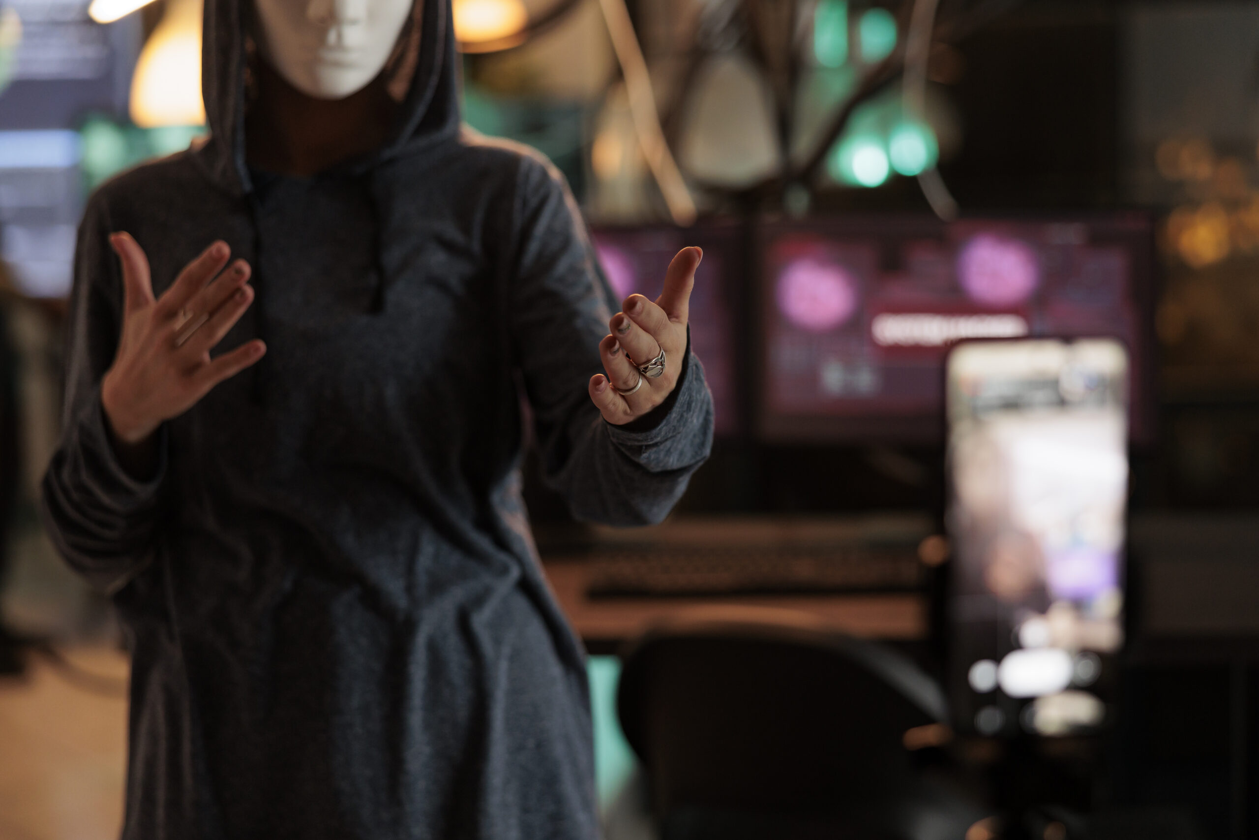 Anonymous figure in hoodie and mask gesturing in front of computer screens.