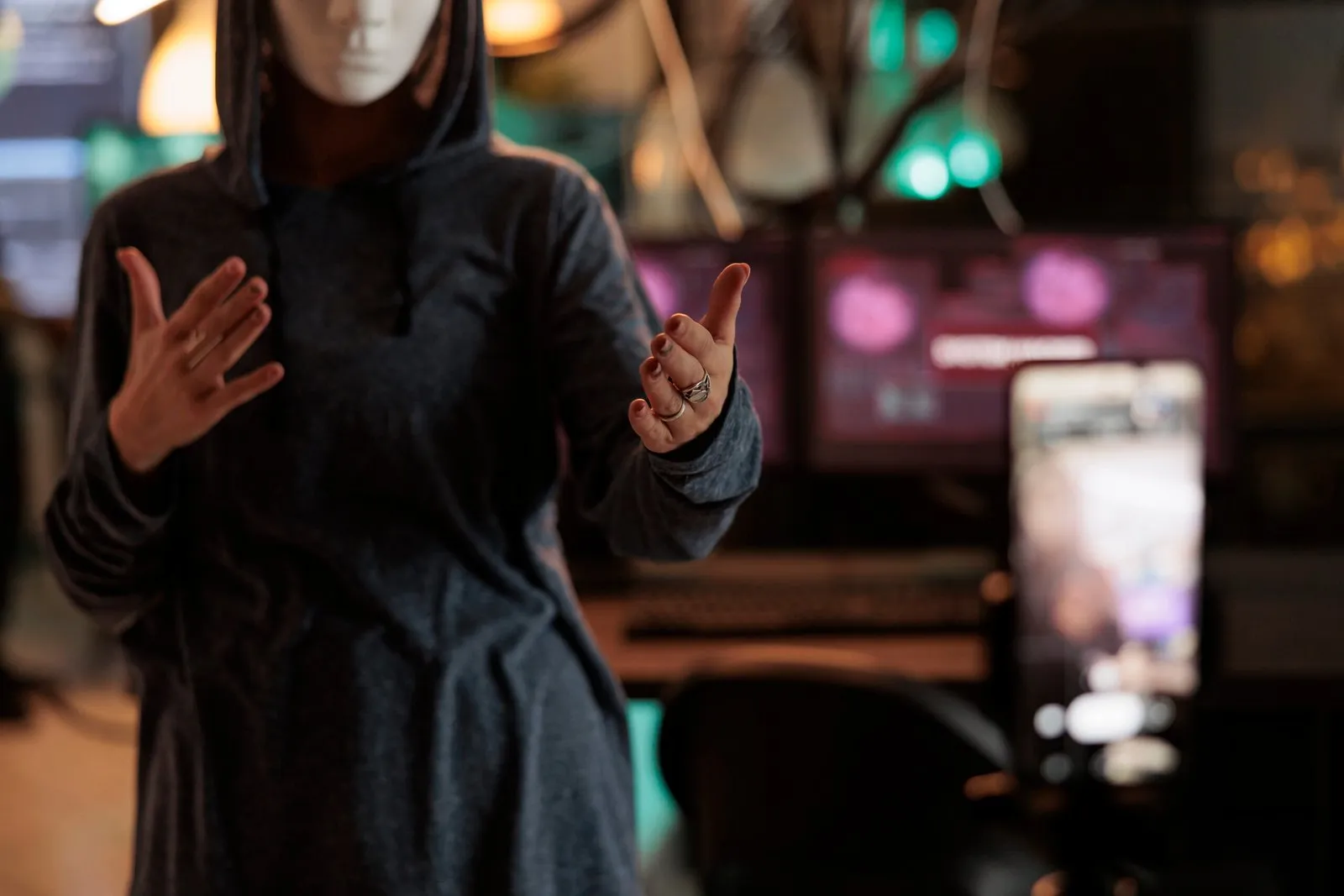 Anonymous figure in hoodie and mask gesturing in front of computer screens.