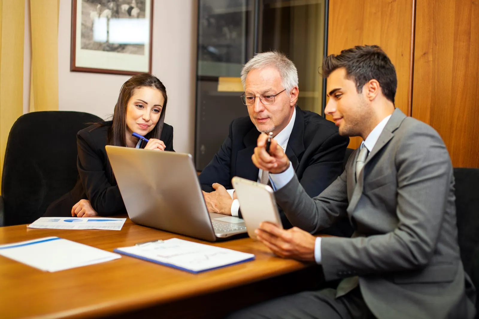 Three professionals in a meeting, reviewing documents and a laptop on a wooden table.