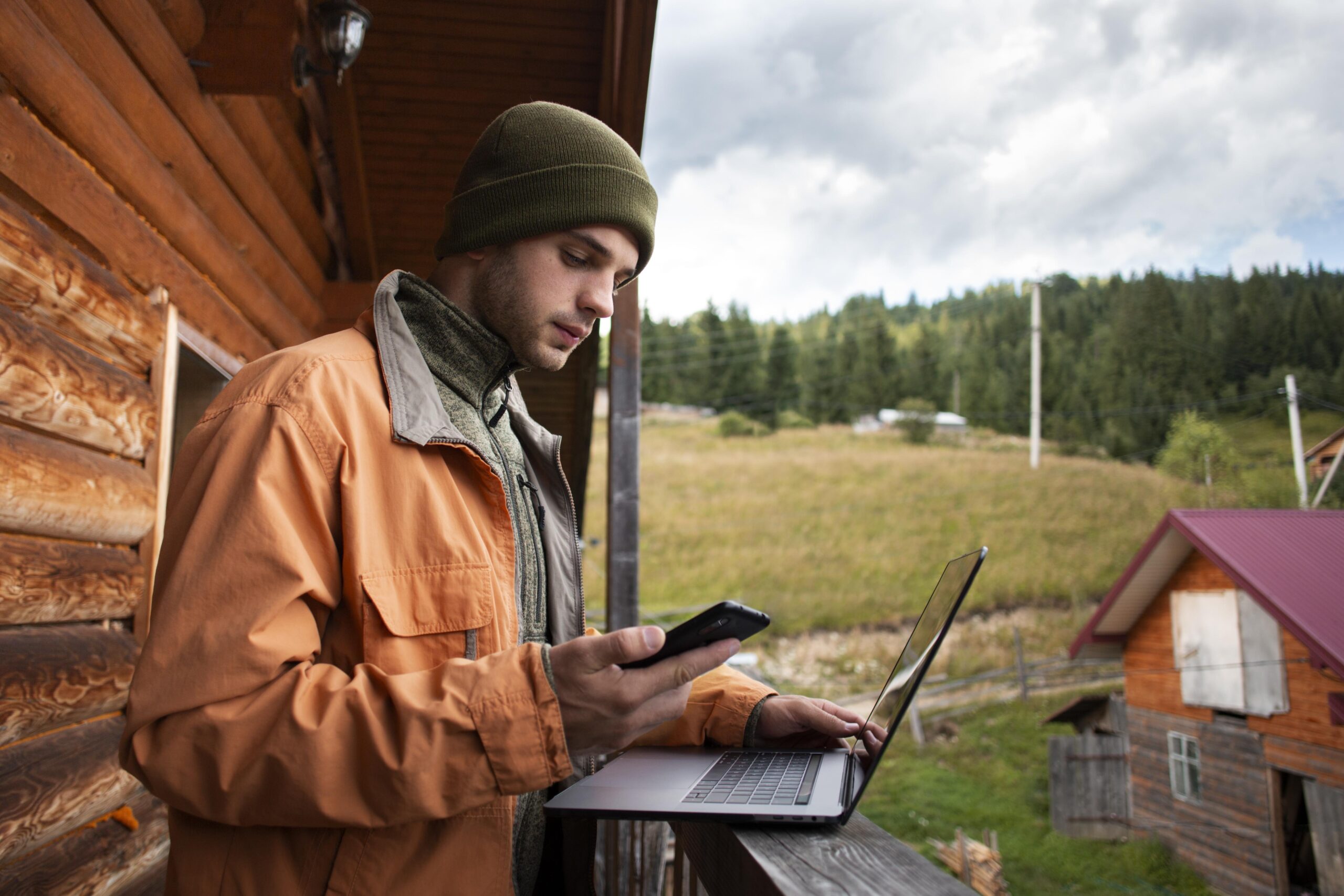 Man on cabin balcony using phone and laptop, working remotely with mountain view.