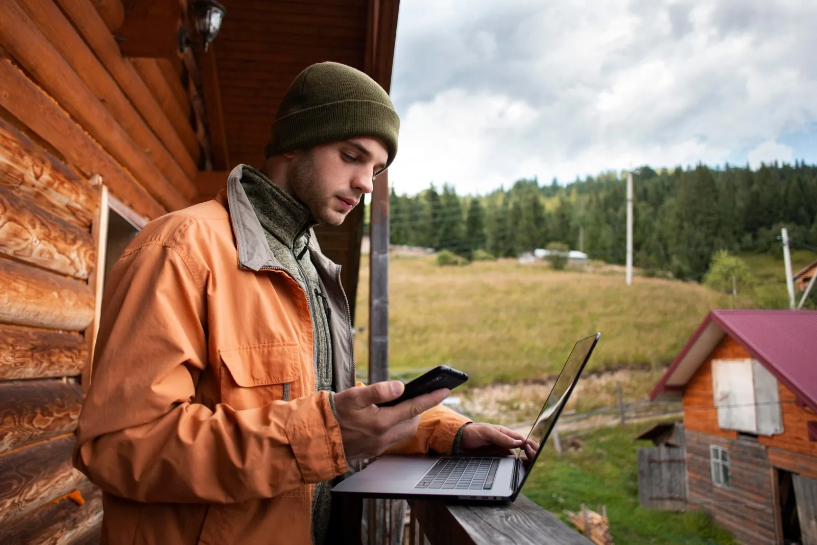 Man on cabin balcony using phone and laptop, working remotely with mountain view.