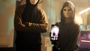 Two masked figures in hoodies in front of a screen displaying 