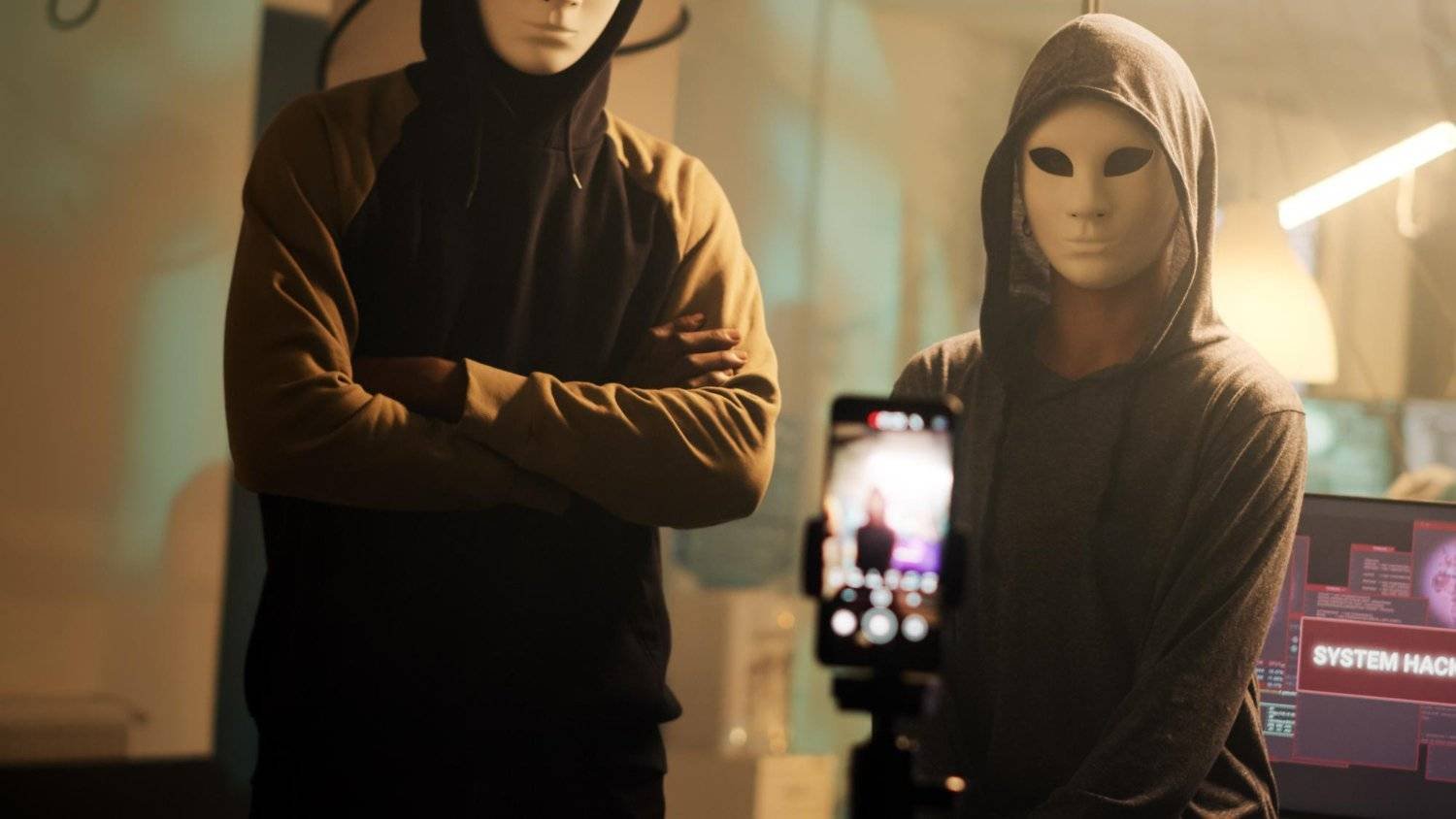 Two masked figures in hoodies in front of a screen displaying 