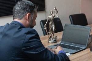Lawyer reviewing documents on laptop with Lady Justice statue on desk.