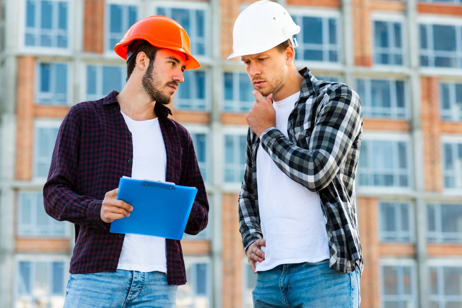 Construction workers reviewing plans on a building site, one in orange, one in white hard hat.