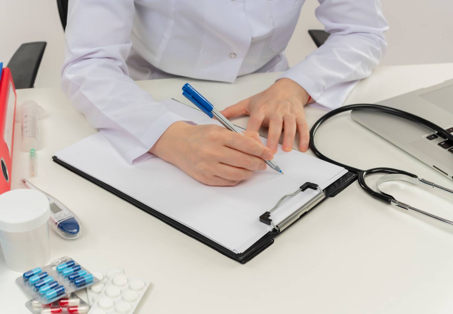 Doctor at desk writing on a clipboard, with medication, stethoscope, and laptop nearby.
